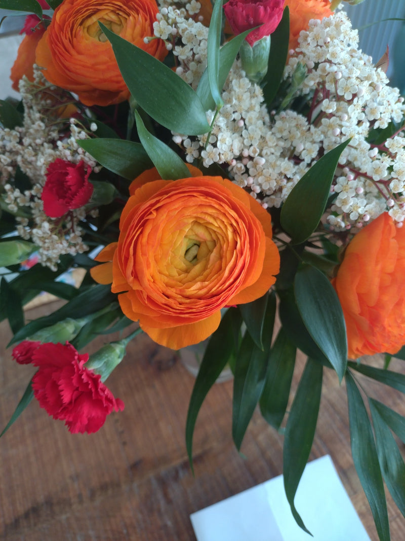 Zu sehen ist eine prachtvolle orangefarbene Ranunkelblüte in einem bunten Blumenstrauß auf einer Holzoberfläche. Die dichten Blütenblätter sind detailreich u...