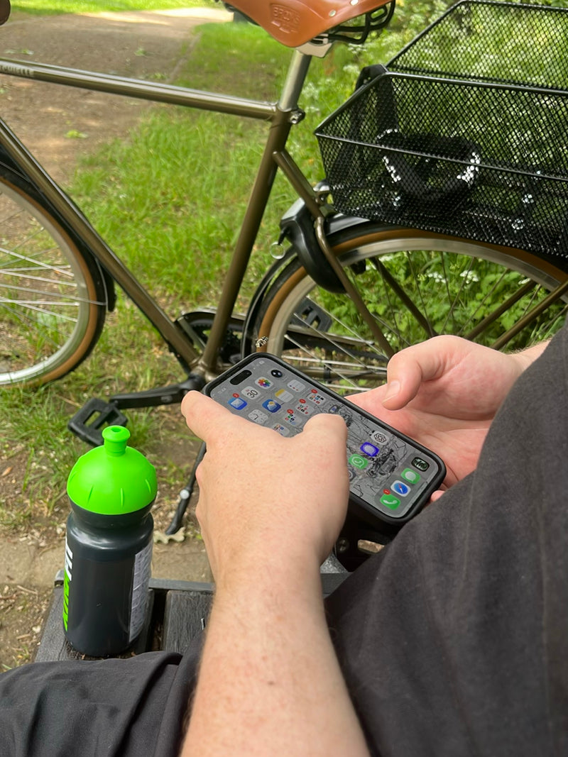 Eine Furchenbiene fliegt im Freien vor einem Smartphone, während eine Person auf einer Parkbank sitzt. Im Hintergrund ist ein Fahrrad zu sehen.