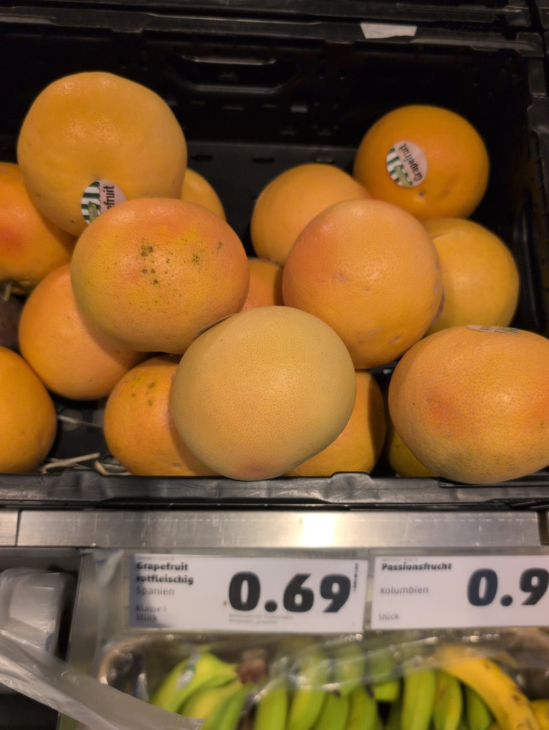 Mehrere reife Grapefruits liegen dicht gedrängt in einer schwarzen Kunststoffkiste im Supermarkt. Auf einigen Früchten sind kleine Aufkleber zu erkennen.
