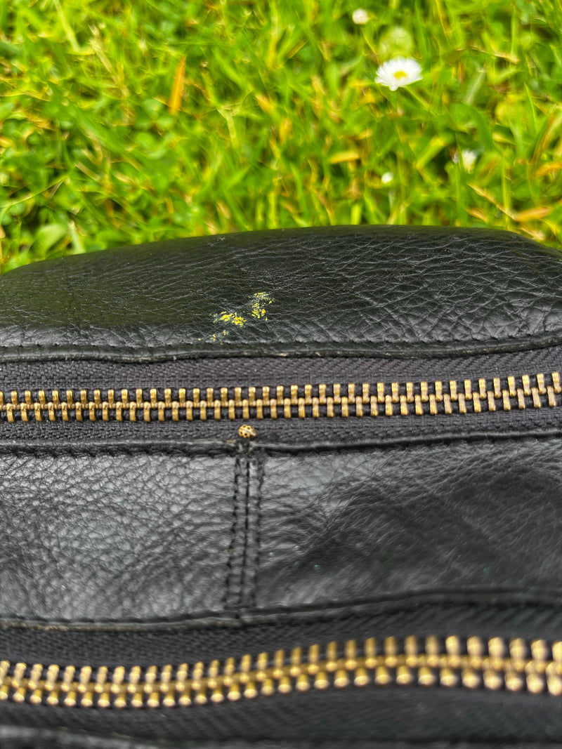 Ein sehr kleiner Sechzehnfleckiger Marienkäfer sitzt auf einer schwarzen Ledertasche nahe am Reißverschluss. Im Hintergrund ist unscharf eine grüne Wiese zu ...